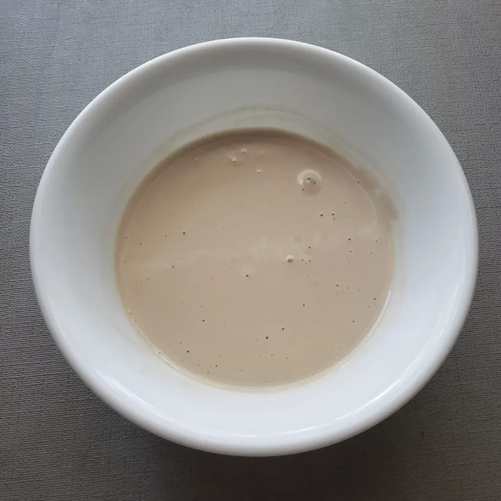 A bowl of creamy, smooth homemade tahini sauce. The texture is rich and velvety, Light background.