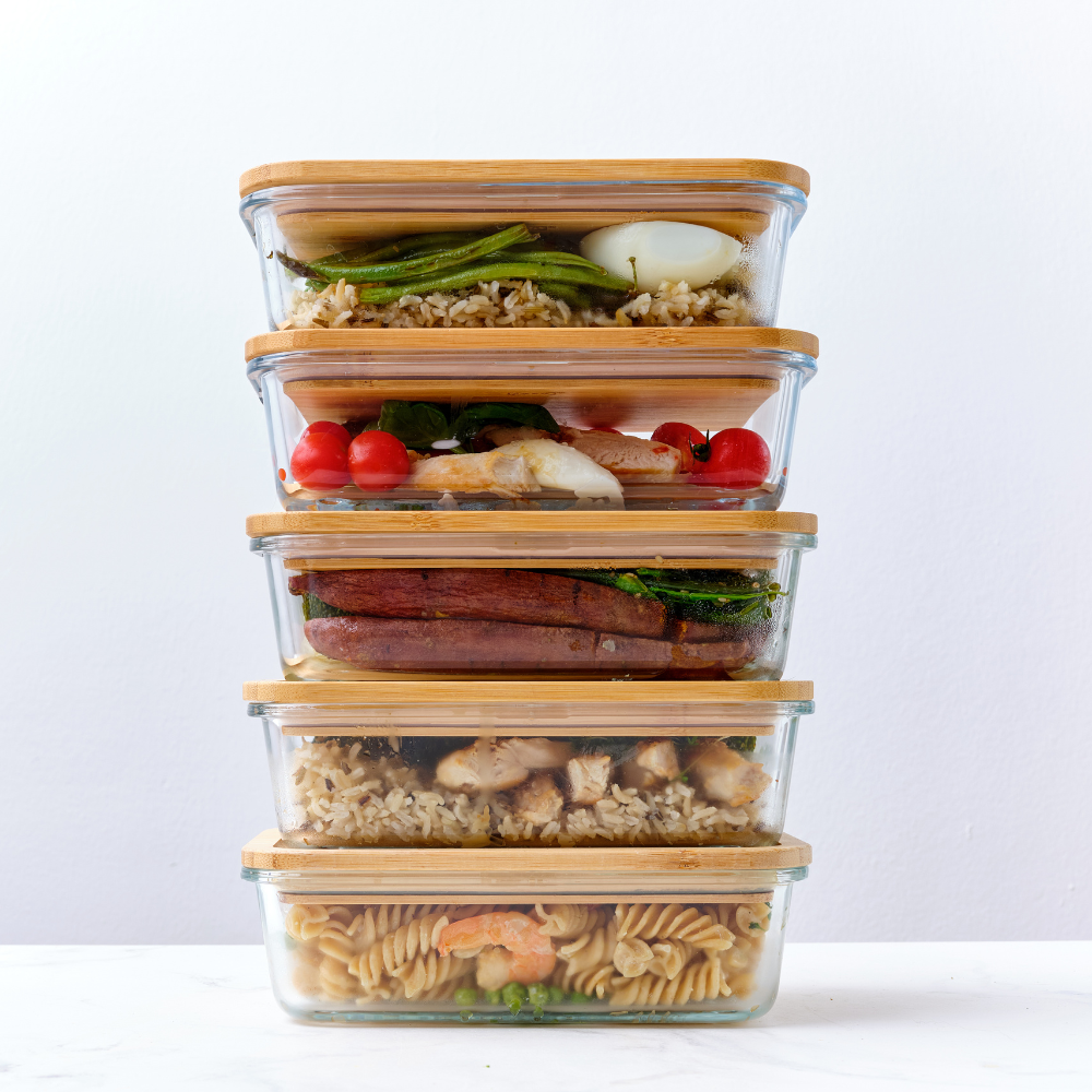 A vertical stack of five rectangular glass meal prep containers with bamboo lids, filled with distinct layers of colorful food including roasted vegetables, a fresh salad, and chicken with rice, resting on a white kitchen counter.
